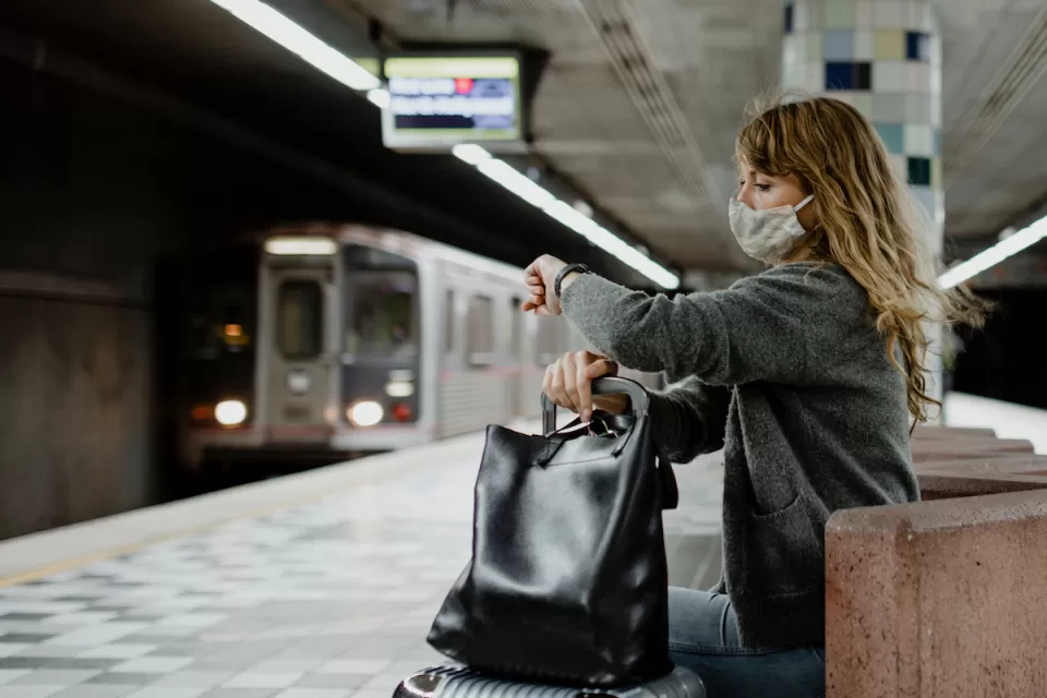 Woman with long hair wearing Covid mask sitting on underground train station bench looking at her watch as subway train approaches in background at Hollywood/Western station in Los Angeles, California.